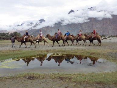 Sechs Personen reiten auf Kamelen in einer malerischen Landschaft mit Bergen und Wolken im Hintergrund.
