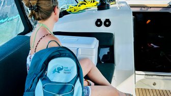 Eine Frau sitzt auf einem Boot und blickt auf das Wasser, während sie einen blauen Rucksack neben sich hat.