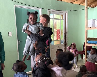 Ein fröhliches Kind wird von einem älteren Jungen in einem hellen Raum hochgehoben, während andere Kinder um sie herum stehen.