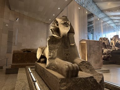 Eine monumentale Sphinx aus Stein in einem modernen Museum, umgeben von weiteren antiken Artefakten.