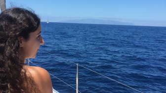 Eine Frau mit lockigem Haar sitzt entspannt auf einem Boot und blickt auf das weite, blaue Meer unter einem klaren Himmel.