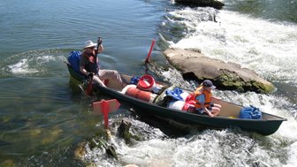 Zwei Kanufahrer navigieren durch eine Stromschnelle, umgeben von klarem Wasser und grüner Vegetation.