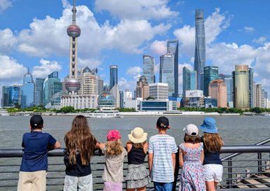 Eine Gruppe von Kindern steht am Wasser und bewundert die beeindruckende Skyline von Shanghai unter einem blauen Himmel.