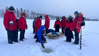 Eine Gruppe von Menschen in roten Jacken steht um ein Loch im Eis, während einer von ihnen mit einem Löffel arbeitet.