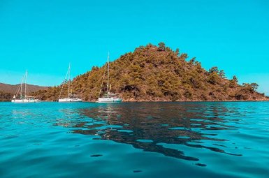 Drei Segelboote liegen ruhig im glitzernden Wasser, umgeben von einer bewaldeten Insel unter strahlend blauem Himmel.