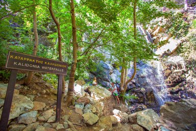 Ein Schild mit der Aufschrift 'Kalidonia Wasserfälle' steht vor einer üppigen, grünen Landschaft mit einem Wasserfall im Hintergrund.