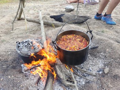 Ein großer Kochtopf mit buntem Eintopf kocht über einem offenen Feuer, umgeben von Sand und Holzstücken.