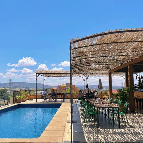 Ein sonniger Poolbereich mit einem Restaurant im Freien, umgeben von Pflanzen und Blick auf die Berge im Hintergrund.