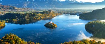 Ein malerischer Blick auf den Bleder See, umgeben von buntem Herbstlaub und majestätischen Bergen im Hintergrund.