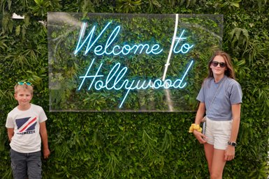 Ein Junge mit blauen Sonnenbrillen und ein Mädchen stehen vor einem leuchtenden 'Welcome to Hollywood'-Schild, umgeben von Pflanzen.