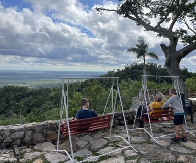 Drei Personen sitzen auf roten Schaukeln und genießen die Aussicht auf eine grüne Landschaft unter einem bewölkten Himmel.