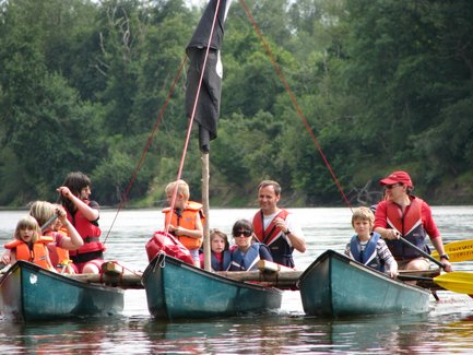 Eine Gruppe von Kindern und Erwachsenen in Kanus, die auf einem ruhigen Fluss paddeln, umgeben von üppigem Grün.
