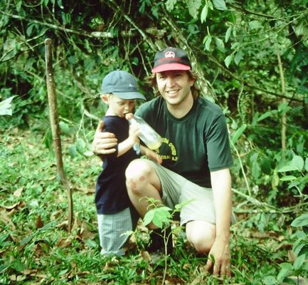 Ein Mann und ein kleiner Junge stehen in einem dichten, grünen Wald, umgeben von üppiger Vegetation und Laub.