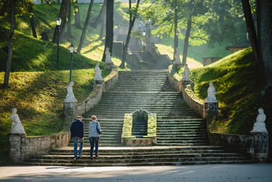 Ein Paar geht eine steinerne Treppe hinauf, umgeben von grünen Wiesen und Statuen, die den Weg säumen.
