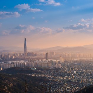 Die Skyline von Seoul bei Sonnenuntergang, mit dem markanten Lotte World Tower, umgeben von sanften Hügeln und Wolken.
