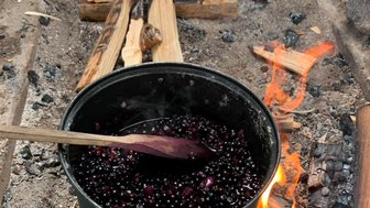 Ein schwarzer Topf mit dunklen Beeren kocht über einem offenen Feuer, umgeben von brennendem Holz und Asche.