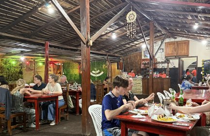 In einem rustikalen Restaurant genießen Gäste an Holztischen ein gemeinsames Essen unter einem strohgedeckten Dach.