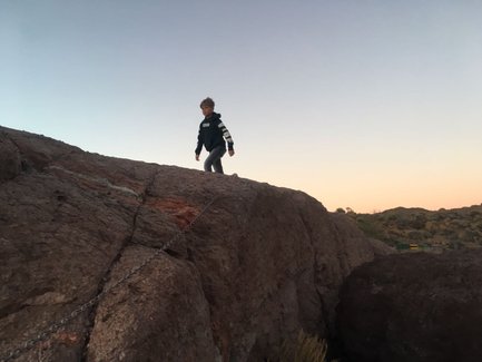 Ein Junge klettert auf einen großen Felsen, während der Himmel in sanften Farben des Sonnenuntergangs leuchtet.