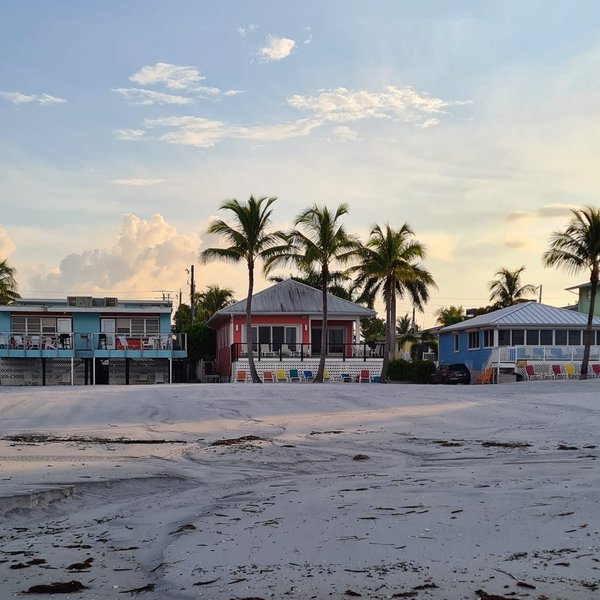 Ein ruhiger Strand mit sanften Wellen, umgeben von Palmen und bunten Häusern im Hintergrund, unter einem klaren Himmel.