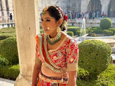 Eine Frau in traditioneller indischer Kleidung lächelt, während sie in einem prächtigen Garten steht, umgeben von historischen Gebäuden.