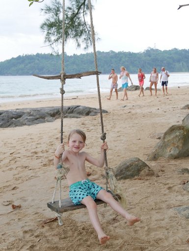 Ein fröhlicher Junge schwingt auf einer Holzschaukel am Strand, umgeben von einer malerischen Küstenlandschaft.