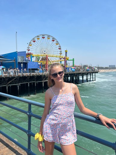 Ein Mädchen steht am Santa Monica Pier, lächelnd, mit einer bunten Achterbahn und dem Ozean im Hintergrund.