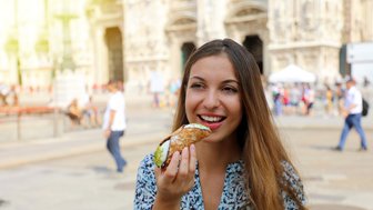Eine junge Frau genießt einen Cannolo in einer belebten Stadtstraße, umgeben von historischen Gebäuden und Passanten.