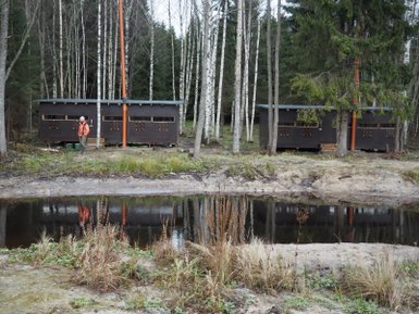 Zwei Beobachtungshütten stehen am Ufer eines ruhigen Gewässers, umgeben von Bäumen und frischem Grün.