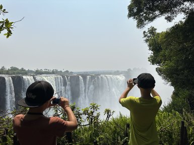 Zwei Kinder stehen am Rand eines Wasserfalls und fotografieren die beeindruckende Aussicht auf die tosenden Wassermassen.