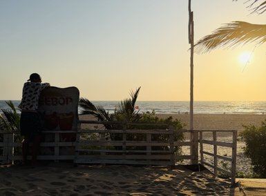 Ein Mann lehnt an einem Schild und blickt auf den Sonnenuntergang über dem Meer, umgeben von Palmen und Sand.