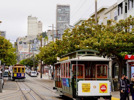 Eine Straßenbahn fährt durch eine belebte Straße in San Francisco, umgeben von hohen Gebäuden und üppigem Grün.