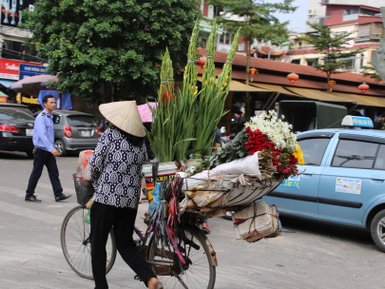 Eine Frau mit einem traditionellen conical Hut fährt mit ihrem Fahrrad, beladen mit bunten Blumen, durch eine belebte Straße.