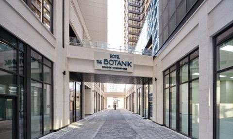 Ein moderner Hotel-Eingang mit dem Schriftzug 'Hotel THE BOTANIK' über einem geräumigen Durchgang, umgeben von hohen Gebäuden.