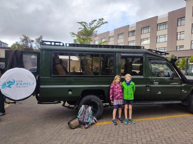 Zwei Kinder stehen lächelnd neben einem grünen Jeep, bereit für ein Abenteuer in Nairobi, mit einem Hotel im Hintergrund.