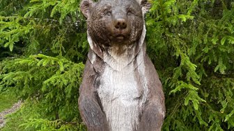 Eine detailreiche Holzskulptur eines Bären steht vor einem grünen Hintergrund aus Bäumen und Sträuchern.