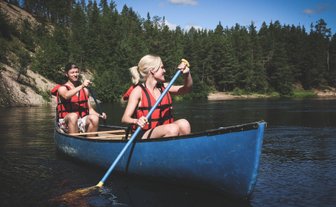 Ein Paar paddelt fröhlich in einem blauen Kanu auf einem ruhigen Fluss, umgeben von üppigem Wald und klarem Himmel.