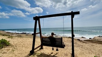 Eine Person sitzt auf einer Schaukel am Strand, umgeben von sanften Wellen und einem klaren, blauen Himmel.