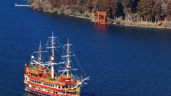 Ein buntes Schiff fährt auf dem ruhigen Wasser eines Sees, umgeben von Bergen und dem majestätischen Fuji im Hintergrund.