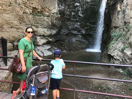Eine Frau in einem grünen Kleid steht neben einem Kind, das an einem Geländer auf einen Wasserfall blickt.