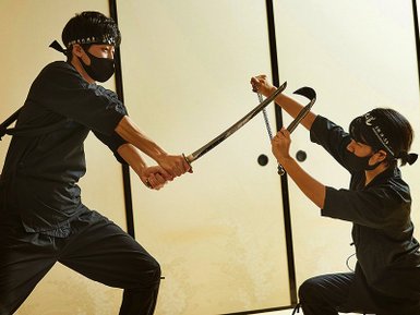 Zwei Kämpfer in traditioneller Ninja-Kleidung führen einen intensiven Schwertkampf in einem stilvollen Raum durch.