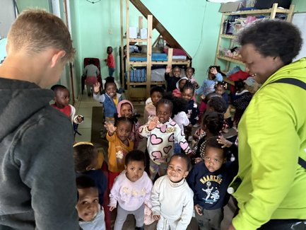 Eine Gruppe von fröhlichen Kleinkindern steht in einem hellen Raum, während ein Erwachsener ihnen freundlich zuwinkt.