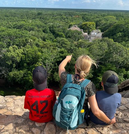 Drei Personen sitzen auf einer Mauer und blicken auf eine grüne Landschaft mit alten Ruinen im Hintergrund.