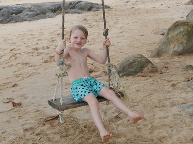 Ein fröhlicher Junge schwingt auf einer Holzschaukel am Strand, umgeben von einer malerischen Küstenlandschaft.