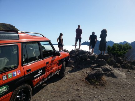 Ein orangefarbener Jeep steht neben einer Gruppe von vier Personen, die auf einer Klippe die Aussicht genießen.