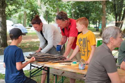 Eine Gruppe von vier Personen arbeitet an einem Tisch im Freien, umgeben von Bäumen und Zelten, während sie Holzstücke sortieren.