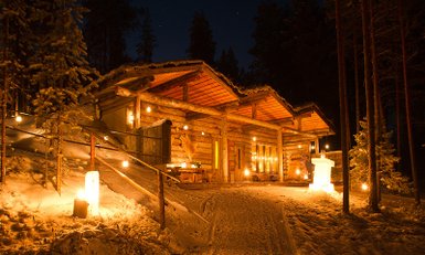 Eine gemütliche Holzhütte im Winterwald, beleuchtet von warmen Lichtern, umgeben von schneebedeckten Bäumen.