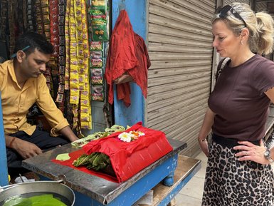 Eine Frau mit einem Leopardenrock betrachtet neugierig ein rotes Tuch mit Blumen auf einem Tisch in einem indischen Straßenladen.