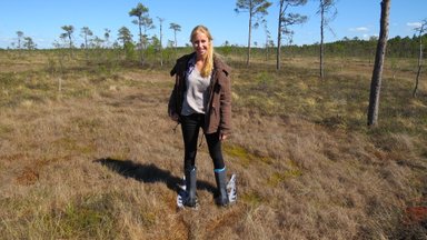 Eine Frau steht in einem Moorgebiet, trägt Gummistiefel und lächelt, umgeben von Bäumen und trockenem Gras.