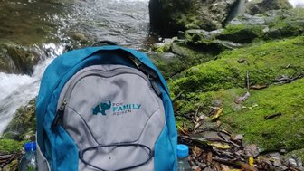 Ein blauer Rucksack steht auf dem feuchten Boden neben einem plätschernden Fluss, umgeben von üppigem Grün und Felsen.