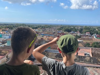 Zwei Jungen stehen an einem Aussichtspunkt und blicken auf die bunten Dächer einer Stadt und das Meer in der Ferne.